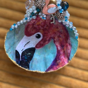 May include: A decorative seashell featuring a flamingo design. The shell is painted with a flamingo portrait in shades of pink, white, and black, set against a blue background. Embellishments include pearls, rhinestones, and a small pink flamingo charm.