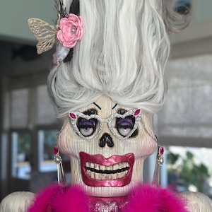 May include: A decorative skull figure with tall, voluminous white hair styled upward, adorned with a pink fabric flower and a beige butterfly. The figure wears white cat-eye glasses with pink gems, long dangling earrings, and a bright pink feather boa.