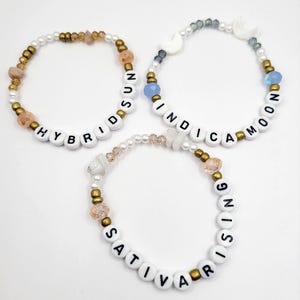 May include: Three beaded bracelets with white beads, gold beads, and small crystals. The bracelets spell out "Hybrid Sun", "Indica Moon", and "Sativa Rising".