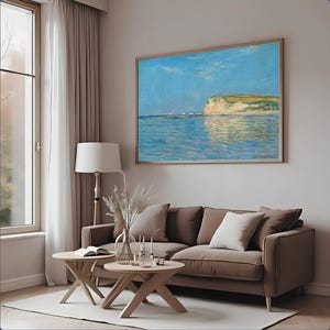 May include: A framed landscape painting depicting a coastal scene with a blue sky and water, and a cliffside. The painting hangs above a brown sofa with white and beige pillows. Two wooden side tables and a floor lamp are in front of the sofa.