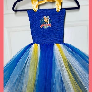 May include: A blue and yellow tulle tutu dress with a blue bodice featuring a Paw Patrol character, Chase, with the text "CHASE" on the chest. The dress has a yellow satin halter strap.