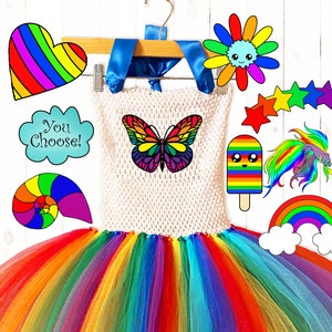 May include: A rainbow tutu with a variety of colorful patches, including a butterfly, a rainbow popsicle, a rainbow fish, a rainbow heart, a rainbow seashell, a rainbow flower, and a rainbow cloud. The text "You Choose!" is on one of the patches.
