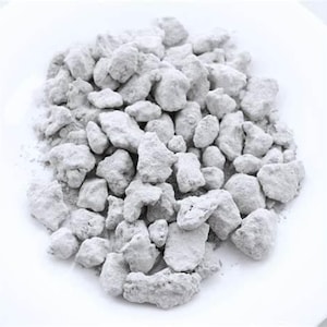May include: A close-up shot of a pile of light gray, irregularly shaped rocks or pebbles. The rocks have a rough, textured surface and appear to be coated in a fine, powdery substance. The rocks are on a white surface.