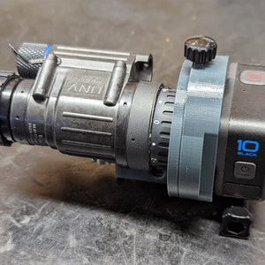 May include: A black and gray night vision scope attached to a black GoPro camera with a gray 3D printed mount. The GoPro camera has a blue button and the text "10 BLACK" on the side.