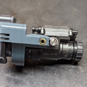 Puede incluir: Una montura gris de impresi&oacute;n 3D unida a un visor de visi&oacute;n nocturna negro. El visor tiene una copa ocular de goma negra y una perilla de ajuste negra.