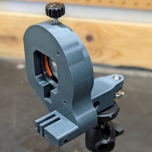 Puede incluir: Un soporte de c&aacute;mara impreso en 3D con una perilla negra y un soporte de tr&iacute;pode plateado. El soporte es gris y tiene un peque&ntilde;o anillo naranja alrededor de la abertura.