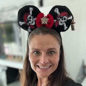 Könnte beinhalten: Eine Frau trägt ein schwarzes Shirt und ein Paar schwarze, gehäkelte Mickey-Mouse-Ohren mit einer roten Schleife und einem goldenen Piraten-Schädel-Anhänger. Die Ohren sind mit einem weißen Totenkopf- und Knochenkreuz-Design verziert.