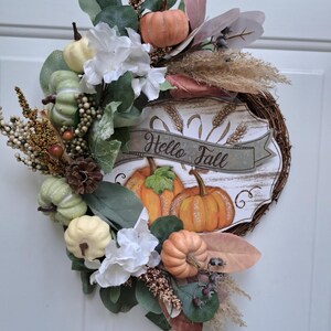 May include: A fall-themed wreath with a rustic design. It features orange and green pumpkins, white flowers, and various foliage. A sign in the center reads "Hello Fall". The wreath is made of woven branches.