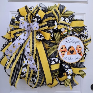 May include: A decorative wreath with a bee theme. The wreath features black and yellow ribbons, polka dots, and daisy patterns. A circular sign reads "Welcome To Our Hive" with bee illustrations. The wreath is designed for a front door.