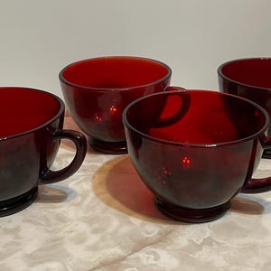 May include: Four ruby red glass teacups with handles. The cups have a rounded bowl shape and sit on a small, circular base. The cups are arranged on a light-colored surface.