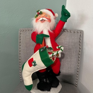 May include: A red and white Santa Claus doll with a white beard and green hat, holding a candy cane and a stocking. The doll is standing on a white base.