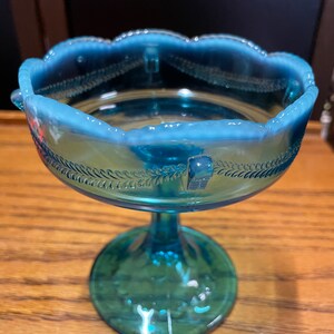 May include: A blue glass pedestal dish with a scalloped rim and a delicate floral design. The dish is made of iridescent glass and has a vintage look.