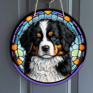 May include: A round stained glass style door hanger featuring a Bernese Mountain Dog puppy with black, white, and brown fur. The dog is looking directly at the viewer with a sweet expression. The stained glass design is colorful and features a variety of shapes and colors.