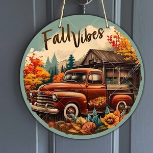 May include: Round wooden sign with the text "Fall Vibes" in brown lettering. The sign features a vintage red truck driving through a forest with fall foliage and a rustic cabin in the background.