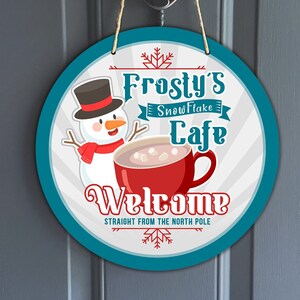 May include: Round, blue and white welcome sign with a snowman and a red mug of hot chocolate. The sign reads "Frosty's Snowflake Cafe Welcome Straight from the North Pole".