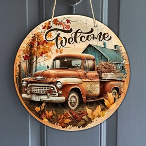 Könnte beinhalten: Ein rundes Holzschild mit einer Willkommens-Botschaft und einem Oldtimer-LKW mit einer Ladefläche voller Heu. Der LKW ist braun und orange mit einer weißen Kabine. Das Schild ist von Herbstblättern in Rot-, Orange- und Gelbtönen umgeben.