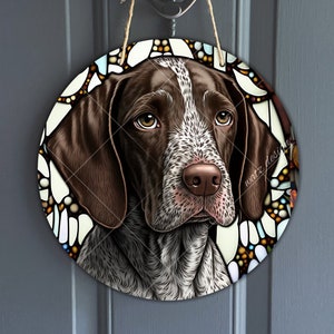 Puede incluir: Cartel de madera redondo con un retrato de un perro Pointer Alemán. El perro tiene pelaje marrón y blanco, con un borde de estilo vidriera. El cartel está colgado en una puerta gris.