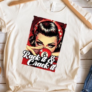 May include: A white t-shirt with a vintage-style graphic of a woman with red hair and a red bandana looking at a pool ball with the text "Rack it & Crack it".