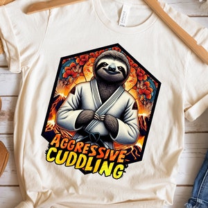 May include: Cream-colored t-shirt featuring a cartoon sloth in a white gi, with the text "AGGRESSIVE CUDDLING." The sloth is set against a fiery background within a black and orange hexagon. The shirt is displayed with sandals and jeans.