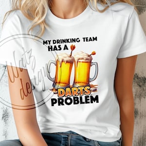 Peut inclure: T-shirt blanc avec un graphique de deux chopes de bière avec des fléchettes qui en sortent. Le texte sur le t-shirt est "My drinking team has a darts problem."