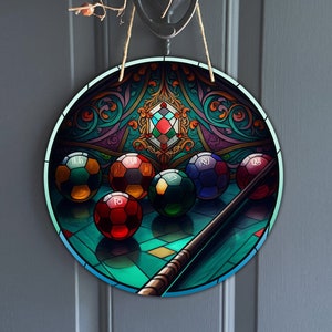 May include: A round, colorful stained glass design featuring a pool table with a cue stick and billiard balls. The balls are numbered 11, 10, 1, 5, 12, and 15.