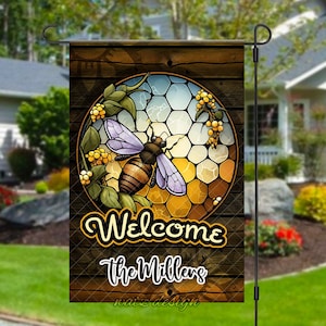 May include: Welcome to the Millers garden flag with a bee and honeycomb design. The flag is brown with a yellow and blue honeycomb pattern and a bee in the center. The text "Welcome the Millers" is printed on the flag.
