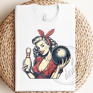 May include: A white t-shirt with a vintage-style illustration of a woman holding a bowling ball and a bowling pin. She is wearing a red dress and a red and white headband. The illustration is in a retro style.