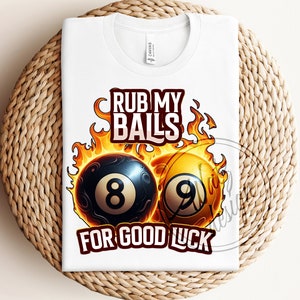 Peut inclure: T-shirt blanc avec un graphique de deux boules de billard en feu. Le texte "Rub My Balls For Good Luck" est imprimé sur le t-shirt.
