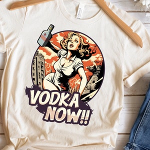 Może przedstawiać: Biały T-shirt z ilustracją w stylu vintage kobiety trzymającej butelkę wódki. Ilustracja jest umieszczona na tle panoramy miasta. Tekst "VODKA NOW!" jest nadrukowany pod ilustracją.