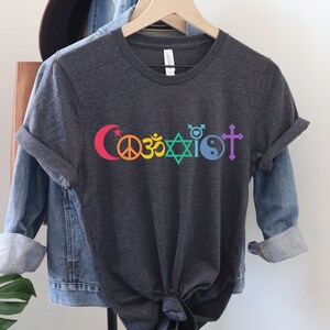 Puede incluir: Una camiseta gris oscuro con la palabra "Coexist" impresa en colores arco&iacute;ris. La palabra est&aacute; compuesta por diferentes s&iacute;mbolos religiosos, incluyendo una luna creciente y una estrella, un signo de paz, un s&iacute;mbolo Om, una estrella de David, un s&iacute;mbolo transg&eacute;nero, un s&iacute;mbolo yin yang y una cruz.