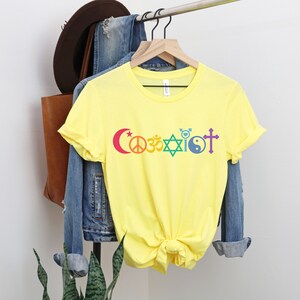 Puede incluir: Camiseta amarilla con la palabra "Coexistir" impresa en colores arco&iacute;ris. La palabra est&aacute; compuesta por diferentes s&iacute;mbolos religiosos, incluyendo una luna creciente, un signo de paz, un s&iacute;mbolo Om, una estrella de David, un s&iacute;mbolo yin-yang y una cruz.