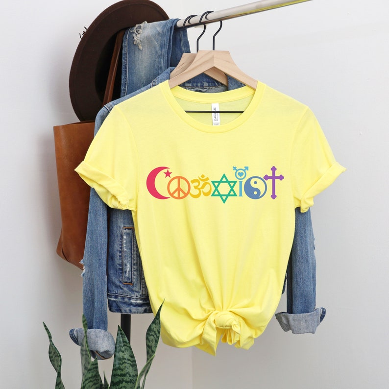 Puede incluir: Camiseta amarilla con la palabra "Coexistir" impresa en colores arco&iacute;ris. La palabra est&aacute; compuesta por diferentes s&iacute;mbolos religiosos, incluyendo una luna creciente, un signo de paz, un s&iacute;mbolo Om, una estrella de David, un s&iacute;mbolo yin-yang y una cruz.