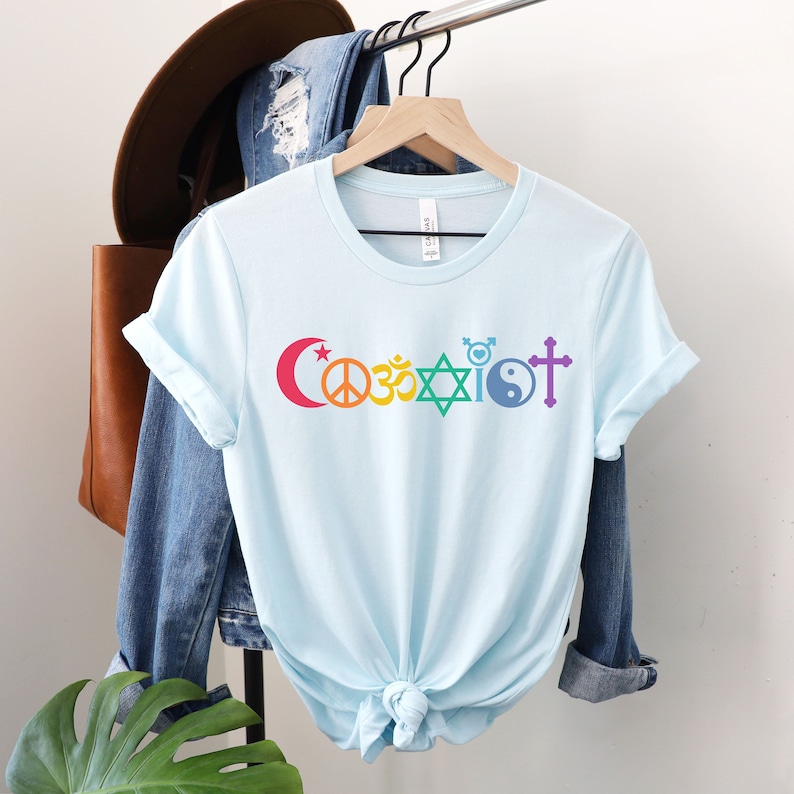 Puede incluir: Camiseta azul claro con la palabra "Coexistir" impresa en colores arco&iacute;ris. La palabra est&aacute; compuesta por diferentes s&iacute;mbolos religiosos, incluyendo una luna creciente, un signo de paz, un s&iacute;mbolo Om, una estrella de David, un s&iacute;mbolo transg&eacute;nero, un s&iacute;mbolo yin-yang y una cruz.