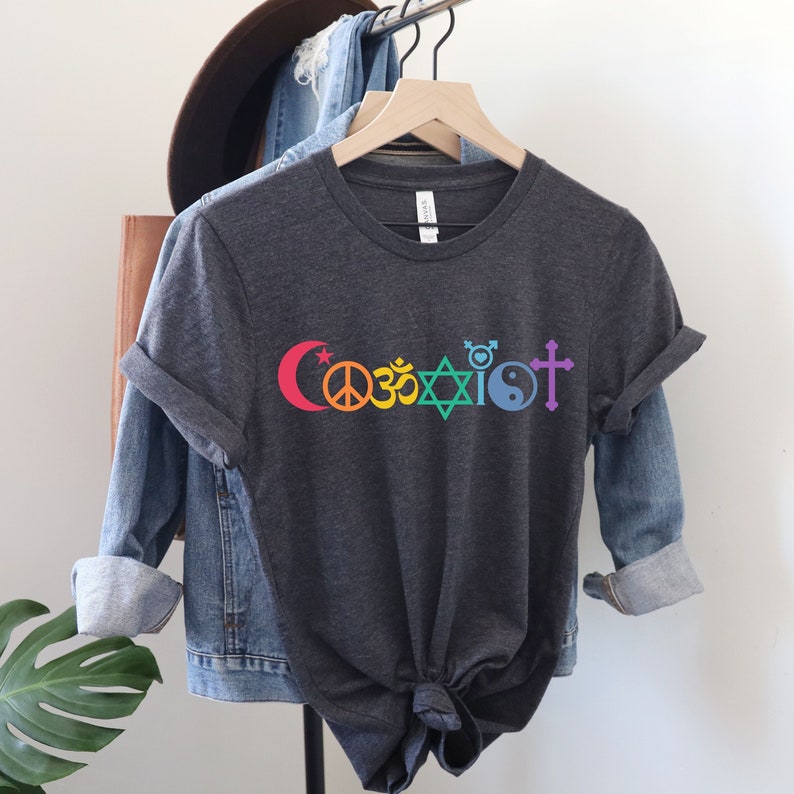 Puede incluir: Una camiseta gris oscuro con la palabra "Coexist" impresa en colores arco&iacute;ris. La palabra est&aacute; compuesta por diferentes s&iacute;mbolos religiosos, incluyendo una luna creciente y una estrella, un signo de paz, un s&iacute;mbolo Om, una estrella de David, un s&iacute;mbolo transg&eacute;nero, un s&iacute;mbolo yin yang y una cruz.