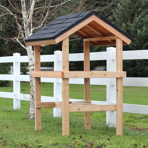 Puede incluir: Un comedero para pájaros de madera con un techo de tejas negras. El comedero tiene dos estantes y se apoya en cuatro patas. El comedero está delante de una valla blanca.