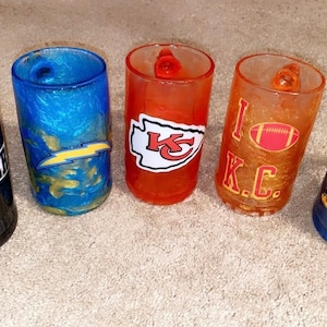 Könnte beinhalten: Fünf Tassen in verschiedenen Farben mit Teamlogos. Die Tassen sind schwarz, blau, orange und gold. Die Logos sind für die Raiders, Chargers, Kansas City Chiefs, Kentucky Wildcats und einen Fußball mit den Buchstaben K.C.