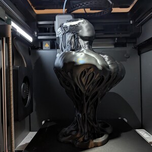 Puede incluir: Un busto negro y brillante del personaje Venom de Marvel Comics, impreso en 3D dentro de una m&aacute;quina. La escultura presenta texturas detalladas y un dise&ntilde;o estilizado, mostrando las caracter&iacute;sticas ic&oacute;nicas del personaje. El busto se exhibe en una impresora 3D.