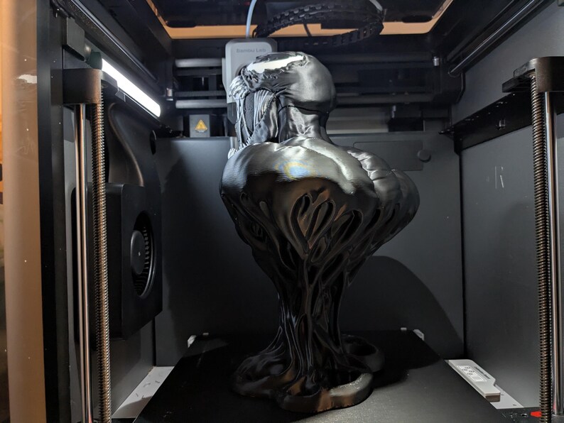 Puede incluir: Un busto negro y brillante del personaje Venom de Marvel Comics, impreso en 3D dentro de una m&aacute;quina. La escultura presenta texturas detalladas y un dise&ntilde;o estilizado, mostrando las caracter&iacute;sticas ic&oacute;nicas del personaje. El busto se exhibe en una impresora 3D.