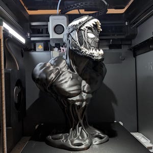 Puede incluir: Un busto en blanco y negro impreso en 3D de Venom, un personaje de Marvel Comics, con un dise&ntilde;o detallado. El busto presenta un torso musculoso y una cabeza con dientes afilados y una m&aacute;scara de Spider-Man. El busto est&aacute; dentro de una impresora 3D.