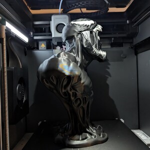 Puede incluir: Un busto negro y plateado impreso en 3D del personaje Venom de Marvel Comics. El busto presenta una escultura detallada de la cabeza y los hombros del personaje, con una expresi&oacute;n amenazante y dientes afilados. El busto est&aacute; dentro de una impresora 3D.