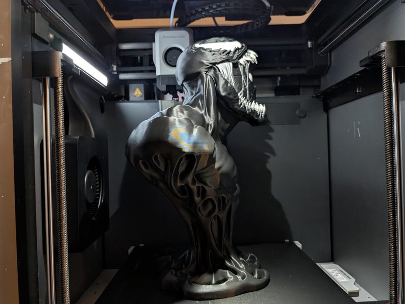 Puede incluir: Un busto negro y plateado impreso en 3D del personaje Venom de Marvel Comics. El busto presenta una escultura detallada de la cabeza y los hombros del personaje, con una expresi&oacute;n amenazante y dientes afilados. El busto est&aacute; dentro de una impresora 3D.