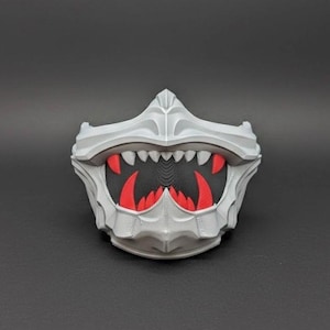 May include: A grey, 3D-printed mask with a fearsome design. The mask has sharp, white teeth and red accents inside the mouth, creating a monster-like appearance. The mask is designed with a detailed, textured surface.