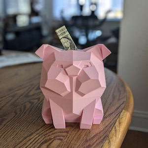 May include: A pink geometric bulldog piggy bank with a dollar bill sticking out of the slot.