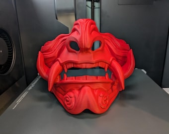 Puede incluir: Una máscara roja impresa en 3D con una expresión feroz. La máscara tiene dientes afilados, una boca ancha y una superficie texturizada.