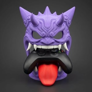 May include: A purple, stylized mask-shaped gaming controller holder. The holder features a demonic face with sharp teeth, horns, and a red tongue. A black gaming controller is held in the mouth of the mask.