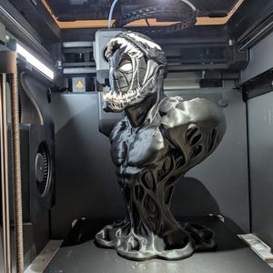 Puede incluir: Un busto en blanco y negro del personaje Venom, con un dise&ntilde;o detallado. El busto est&aacute; en una impresora 3D, mostrando el proceso de impresi&oacute;n. La figura tiene un f&iacute;sico musculoso y una expresi&oacute;n amenazante.