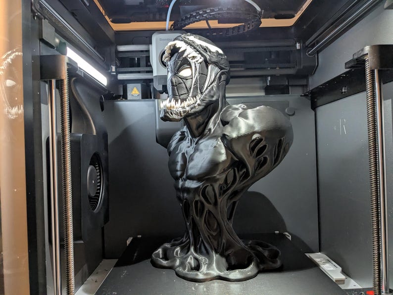 Puede incluir: Un busto en blanco y negro del personaje Venom, con un dise&ntilde;o detallado. El busto est&aacute; en una impresora 3D, mostrando el proceso de impresi&oacute;n. La figura tiene un f&iacute;sico musculoso y una expresi&oacute;n amenazante.