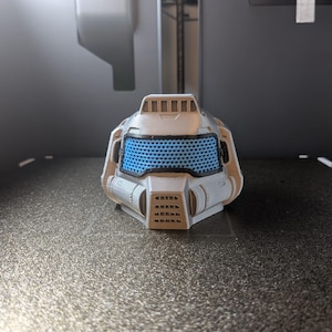 May include: A 3D-printed helmet in grey and blue. The helmet has a blue mesh visor and geometric details. The design suggests a futuristic or sci-fi theme, possibly for cosplay or display.