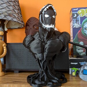 Puede incluir: Busto negro de Venom con detalles blancos en la cabeza y la boca. La estatua tiene un torso musculoso y una base texturizada y goteante. Otros elementos en el fondo incluyen una l&aacute;mpara, una figura de robot y una caja de cereales.