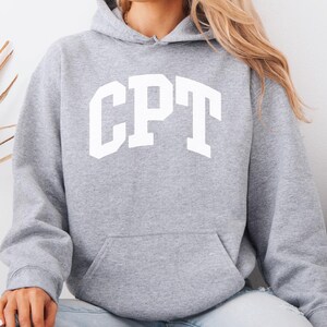 Könnte beinhalten: Ein hellgrauer Kapuzenpullover mit den Buchstaben "CPT" in weißen Blockbuchstaben auf der Brust. Der Kapuzenpullover hat eine Vordertasche und eine Kapuze. Der Text "*SPORT GREY" befindet sich in der oberen rechten Ecke. Das Model trägt hellblaue Jeans.